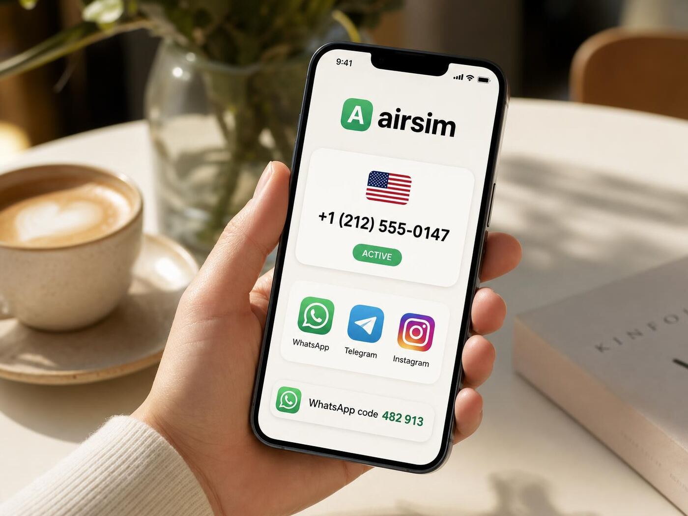 A hand holding a phone at a sunlit cafe table, the Airsim app showing a live US number and a WhatsApp code